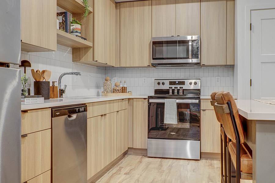 Modern kitchen with light wood cabinets, stainless steel appliances, white tile backsplash, and wood-look flooring.