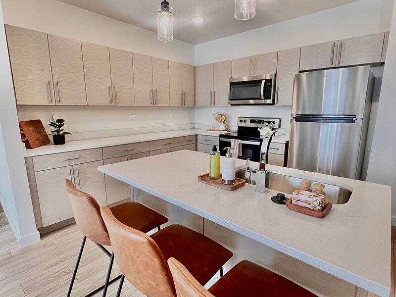 Modern kitchen with light wood cabinets, white quartz counters, stainless steel appliances, large island with sink, and four brown leather bar stools. Minimalist and bright.