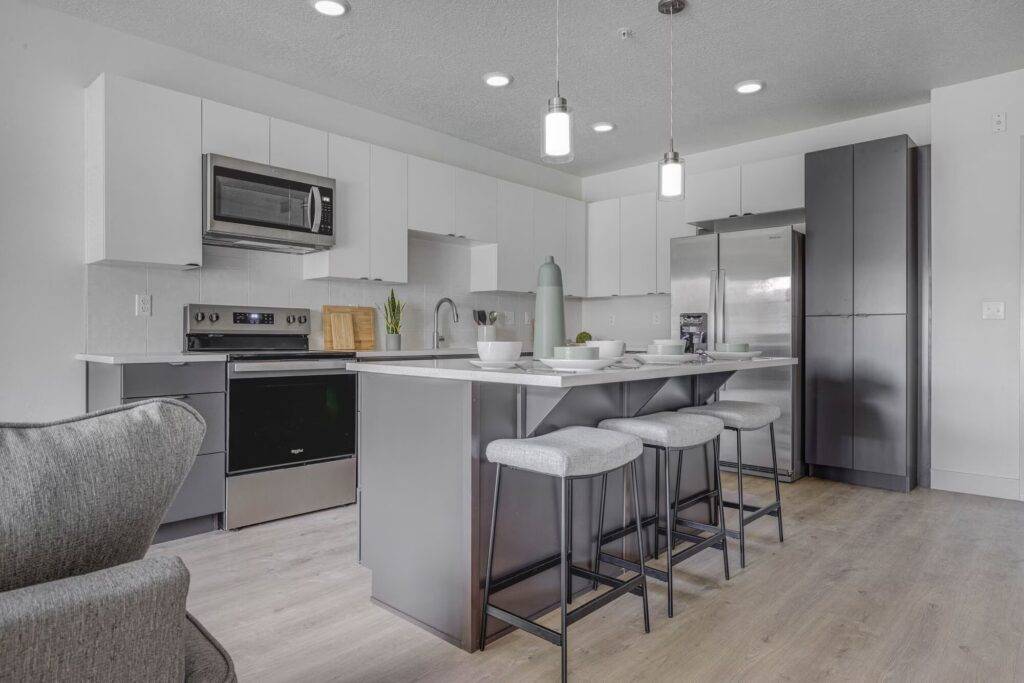 Modern kitchen with white upper cabinets, gray lower cabinets, stainless steel appliances, large island with seating for four, pendant lights, and light wood flooring.