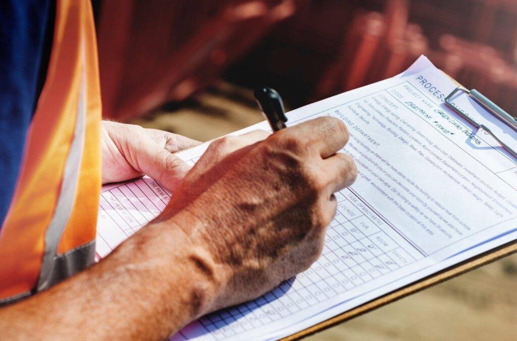 A man wearing an orange vest is writing on a clipboard focused on his task in an outdoor setting